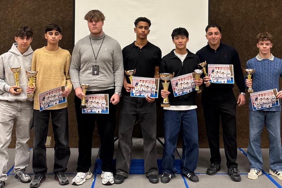 Gruppe von sieben Jugendlichen mit Pokalen und Urkunden vor einer Leinwand in einem Veranstaltungsraum.