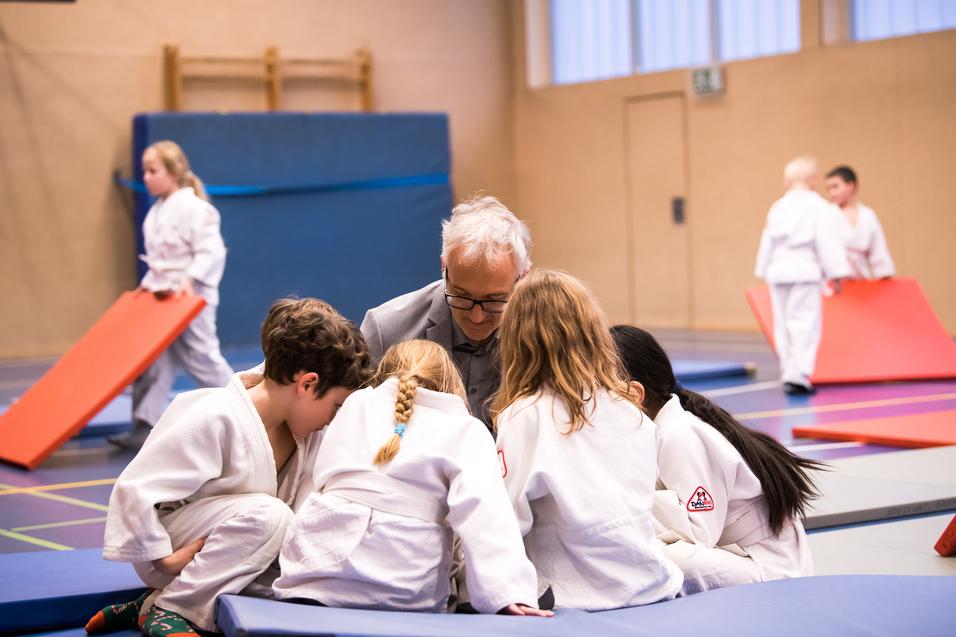 Sechs Kinder in Judogis beugen sich um einen älteren Trainer in einem Sportraum, während andere Kinder im Hintergrund spielen.